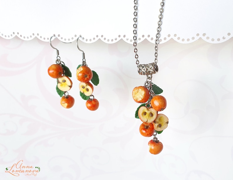 May include: A jewelry set featuring a necklace and earrings with a fruit theme. The necklace has a silver chain with a pendant of miniature orange apples, some sliced to reveal the interior. The earrings mirror the design, with green leaves.