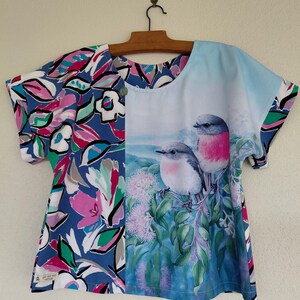 May include: A short-sleeved top featuring a split design. The left side displays a vibrant floral pattern in blue, pink, white, and green. The right side depicts two small birds with pink chests perched on green branches against a light blue sky.