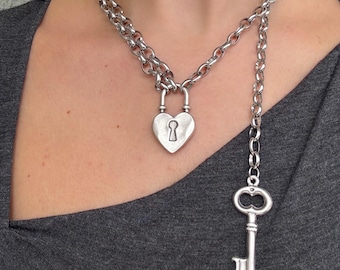 May include: Silver-coloured necklace with a heart-shaped padlock pendant and a dangling key. The necklace is made of a chunky chain and is designed to be worn around the neck. The key hangs down from the chain.
