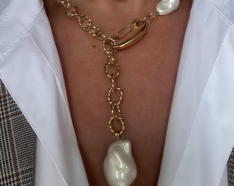 May include: Two-strand necklace featuring large, irregular white pearls and gold-tone chains. The necklace has a unique design with a large pearl pendant and a gold clasp. The jewelry is worn over a white collared shirt.