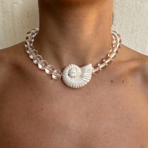 May include: A clear beaded necklace featuring a large, white, seashell-shaped pendant. The round, transparent beads encircle the neck, with the pendant displaying a detailed spiral design. The necklace is a statement piece.