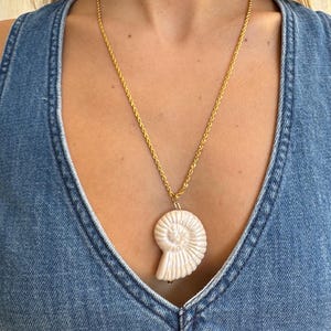 May include: A gold-colored chain necklace with a large, off-white, seashell-shaped pendant. The pendant has detailed spiral ridges. The necklace is worn over a denim top.
