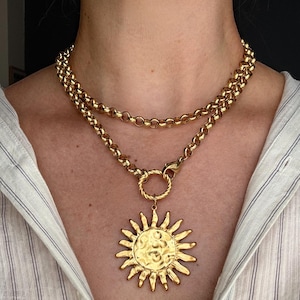 May include: A gold-toned necklace featuring a double-layered rolo chain and a large, circular sun pendant with a textured, hammered center and radiating rays. The pendant is attached to the chain by a twisted circular ring and a lobster clasp.