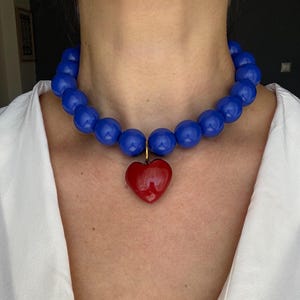 Puede incluir: Un collar tipo gargantilla que presenta una sola hilera de cuentas redondas, grandes y brillantes de color azul. Un colgante rojo liso en forma de corazón cuelga del centro de la hilera de cuentas mediante un pequeño anillo de metal dorado.