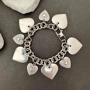 Può includere: Bracciale con ciondoli color argento con ciondoli a forma di cuore. Il bracciale ha un design a catena grossa e una chiusura. Alcuni ciondoli a forma di cuore sono lisci, altri hanno un motivo inciso. Il bracciale è esposto su una superficie grigio scuro. Lunghezza bracciale: circa 19 cm.