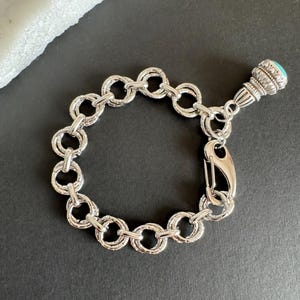 Könnte beinhalten: Ein silberfarbenes Kettenarmband aus ineinandergreifenden, kreisförmigen Gliedern mit strukturierter Oberfläche. Das Armband verfügt über einen großen Karabinerverschluss und einen dekorativen Anhänger mit einem kleinen, runden, blauen Stein.