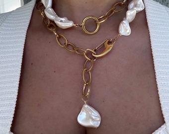 May include: A two-strand necklace featuring a gold chain and large, irregular white pearls. The necklace has a gold clasp and a single pearl pendant. The jewelry is worn over a white sweater.