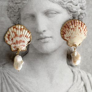Può includere: Orecchini a conchiglia con bordatura dorata e perla barocca pendente. Gli orecchini presentano un motivo naturale a conchiglia con strisce rosse e bianche. Lo sfondo è un'immagine in scala di grigi di una statua classica.