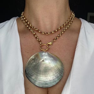 Puede incluir: Un collar de cadena rolo doble en tono dorado con un gran colgante circular de nácar iridiscente. El colgante está sujeto a la cadena mediante una anilla dorada trenzada y un cierre de mosquetón.