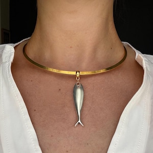 May include: A gold-toned metal choker necklace featuring a smooth, polished silver-toned fish pendant suspended from a central gold-toned bail. The necklace sits close to the base of the neck, showcasing a minimalist design with a metallic finish.