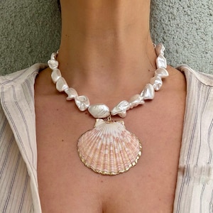 May include: A statement necklace featuring large, irregular white pearls. The pendant is a large, pink and white seashell with a gold-colored trim. The necklace is worn against a striped shirt.