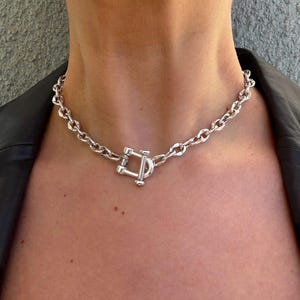 May include: A silver-toned chain necklace with a large, decorative clasp. The necklace is made of thick, rounded links and is worn around the neck. The clasp is a rectangular shape with a bar through the center. The necklace is a bold statement piece.
