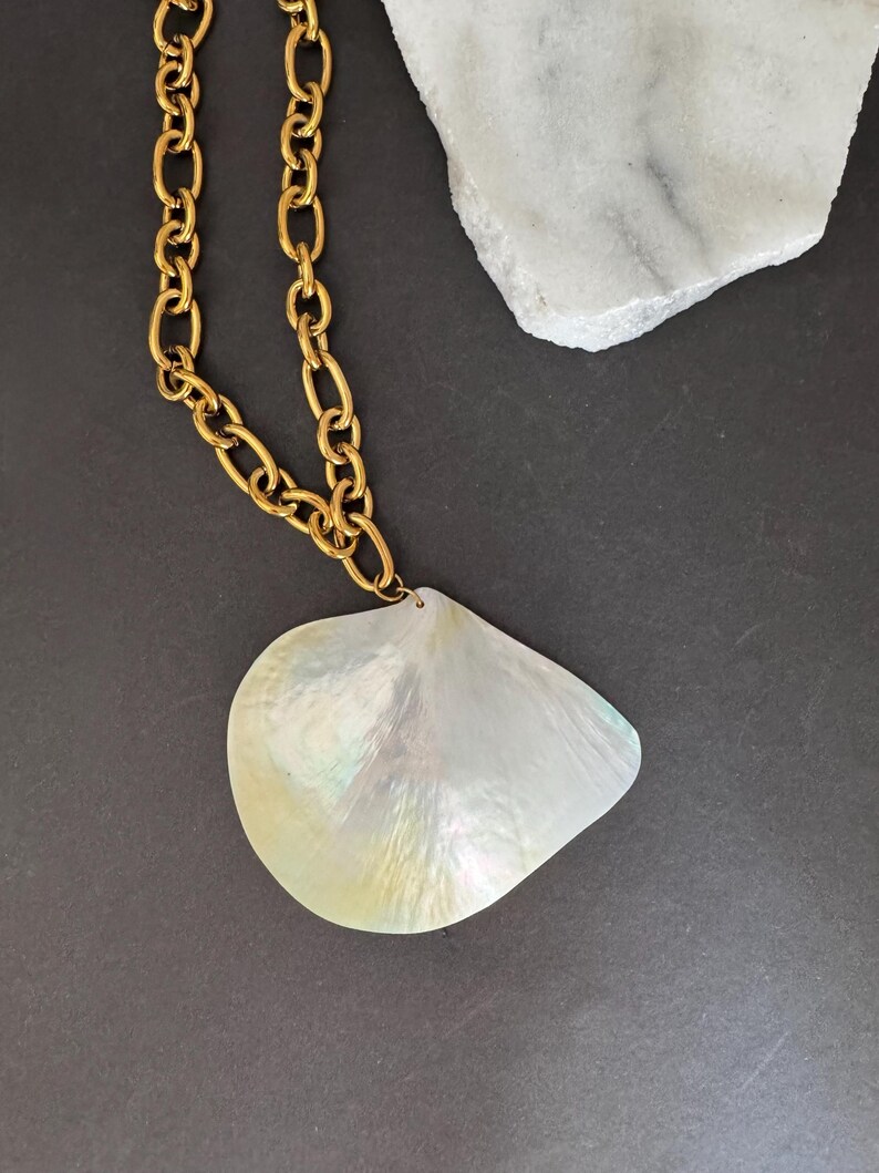 May include: A gold-tone chain necklace with a large, pearlescent, teardrop-shaped pendant. The pendant has iridescent qualities, reflecting a range of colors. The necklace is displayed on a dark gray surface.