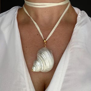 Peut inclure: Un collier composé d'un grand pendentif en coquillage blanc irisé à la texture lisse et spiralée. Le coquillage est fixé à une bélière en métal doré et suspendu à un long ruban en tissu crème fin qui s'enroule autour du cou.