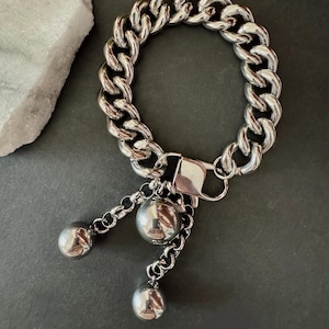 May include: A silver-toned curb chain bracelet featuring a large lobster claw clasp. Three polished silver spheres of varying sizes hang from the clasp, suspended by short lengths of delicate chain, creating a dangling charm effect.