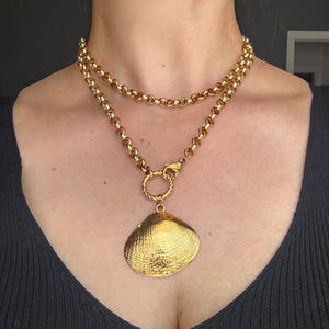 May include: A gold-toned necklace featuring a double-layered rolo chain design. The necklace includes a textured, circular twisted ring connector that holds a large, polished gold-toned seashell pendant with a detailed, ridged surface texture.