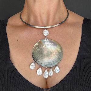 May include: A silver-toned metal choker necklace featuring a large, iridescent, circular shell pendant. The pendant is accented with a single small white pearl at the top and five dangling, irregular white pearls attached to the bottom edge.
