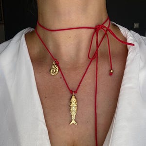 May include: A red cord necklace featuring a gold-toned fish pendant and a smaller gold-toned ammonite shell charm. The cord wraps around the neck and ties in a knot, with two gold-toned beads at the ends of the dangling cord sections.
