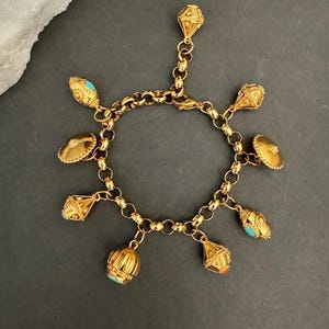 Könnte beinhalten: Ein goldfarbenes Bettelarmband mit einer runden Gliederkette und mehreren hängenden Anhängern. Die Anhänger umfassen texturierte geometrische Formen, kuppelförmige Elemente und glockenartige Formen, teilweise akzentuiert mit kleinen türkisfarbenen und orangefarbenen Steinen.