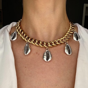 May include: A gold-toned chunky chain necklace featuring five silver-toned cowrie shell charms hanging at intervals along the length of the chain. The necklace rests against the neck, showcasing a high-shine metallic finish on both the chain and shells.