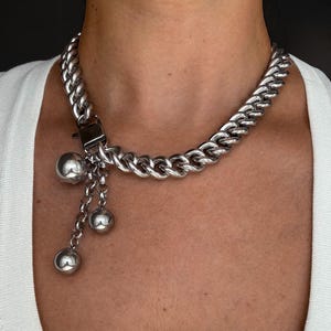 Peut inclure: Un collier à chaîne argentée composé de maillons épais et entrelacés. Le collier présente un fermoir carré auquel sont suspendues trois chaînes fines de longueurs différentes, chacune terminée par une sphère métallique argentée et polie.