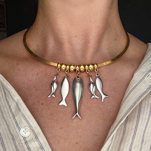 May include: A gold-toned metal choker necklace featuring a series of five silver-toned fish-shaped charms suspended from a row of small, round gold-toned beads. The necklace has a smooth, rigid band design that rests around the base of the neck.