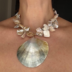 May include: A statement necklace featuring large, iridescent shell pieces in shades of cream, gold, and silver. The necklace has a large, fan-shaped pendant suspended from a strand of smaller, irregular shell fragments, creating a unique and eye-catching design.