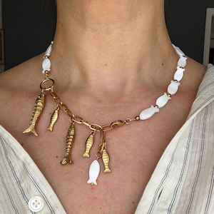 Può includere: Una collana dorata con ciondoli a forma di pesce. La collana presenta una catena con ciondoli a forma di pesce dorati e perline bianche a forma di pesce. È un accessorio di moda.