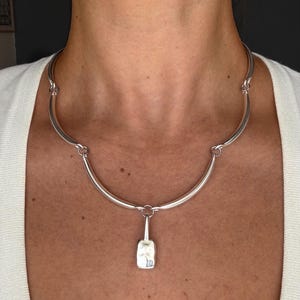 Può includere: Una collana color argento caratterizzata da una serie di segmenti metallici curvi e lisci collegati tra loro. La collana include un ciondolo centrale di forma rettangolare con una superficie martellata e testurizzata, sospeso a un anello circolare.