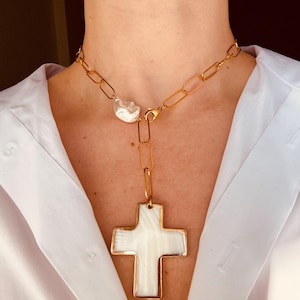 Può includere: Una collana a catena dorata con una grande perla bianca e un ciondolo a forma di croce bianca. La croce è realizzata in un materiale bianco con una cornice dorata.