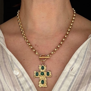 May include: Gold-toned necklace featuring a large cross pendant with green gemstone accents. The chain is a thick, rounded link style with a toggle clasp. The cross design has an ornate, textured appearance.