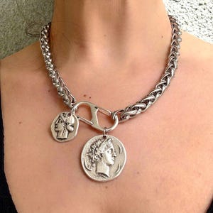 Op de afbeelding: Zilverkleurige ketting met twee muntbedels. De ketting heeft een dik, gevlochten kettingontwerp en een grote sluiting. De muntbedels tonen klassieke figuren in profiel.
