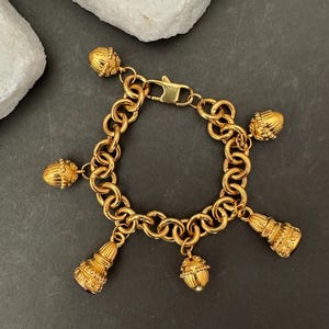 Può includere: Bracciale dorato con ciondoli decorativi. Il bracciale ha un design a catena con chiusura a moschettone. I ciondoli includono pendenti a forma di ghianda e nappa, aggiungendo un tocco di eleganza ai gioielli.