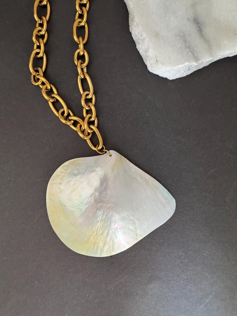 May include: A gold-tone chain necklace with a large, iridescent, teardrop-shaped pendant. The pendant has a pearlescent sheen with hints of green and yellow. The necklace is displayed on a dark gray surface.