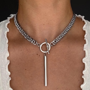 May include: A silver-toned chain necklace featuring a toggle clasp closure at the center. The necklace includes a smooth, cylindrical pendant hanging vertically from the circular ring of the toggle mechanism, creating a clean and metallic aesthetic.