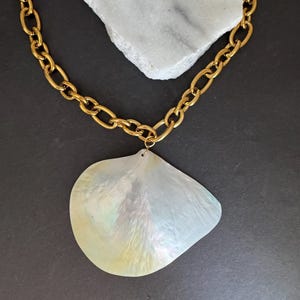 May include: A gold-colored chain necklace with a large, iridescent, pearlescent pendant. The pendant is a shell shape with a subtle, shimmering effect. The necklace is displayed on a white stone against a dark background.