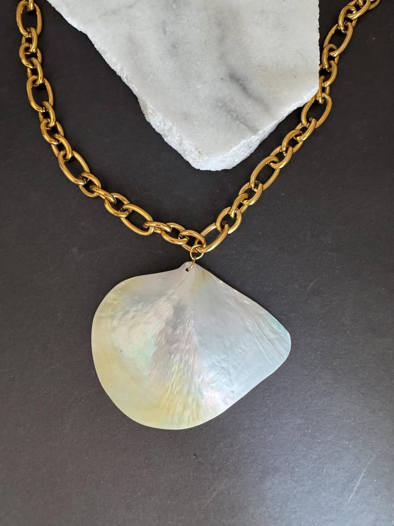May include: A gold-colored chain necklace with a large, iridescent, pearlescent pendant. The pendant is a shell shape with a subtle, shimmering effect. The necklace is displayed on a white stone against a dark background.