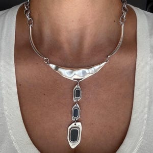 Op de afbeelding: Een zilverkleurige ketting met een gebogen, gehamerd metalen kraagstuk verbonden aan een gedraaide ketting. Een verticale hanger hangt in het midden, bestaande uit drie onregelmatig gevormde zilveren schakels met donkere, rechthoekige centra.