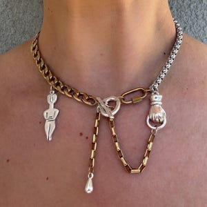 Könnte beinhalten: Halskette aus Mischmetall mit gold- und silberfarbenen Kettengliedern. Die Halskette hat verschiedene Anhänger, darunter eine kleine Figur, eine Faust und einen Tropfenanhänger. Sie wird mit einem Knebelverschluss und einem herzförmigen Verbinder befestigt.
