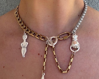 May include: A mixed-metal necklace featuring a gold and silver chain design. The necklace has a variety of charms, including a small figure, a fist, and a teardrop pendant. The necklace is secured with a toggle clasp and a heart-shaped connector.