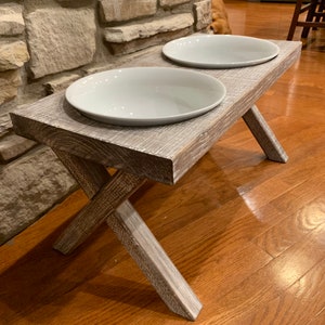 May include: A white wooden dog bowl stand with two white ceramic bowls. The stand has a rustic, distressed finish and a criss-cross base.