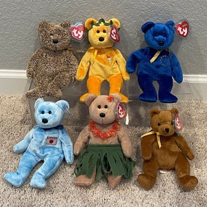 May include: A collection of six beanie baby plush toys. The toys are all different colors and have different designs. The toys are all sitting on a white surface. The toys are all in good condition.