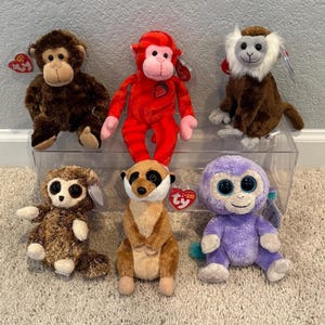 May include: A collection of six plush animal toys including a brown monkey, a red monkey with a strawberry patch, a brown and white monkey, a brown textured monkey, a tan meerkat, and a purple monkey, each featuring a red Ty heart-shaped tag.
