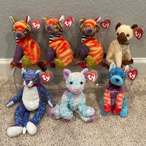 May include: Seven colorful plush cat toys with Ty tags. The cats are in various poses, including sitting, standing, and lying down. The cats are all different colors, including orange, blue, green, and white.