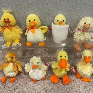 May include: A collection of eight yellow plush duck toys. The ducks are all different sizes and have different features, such as polka dots, bows, and fluffy feathers.