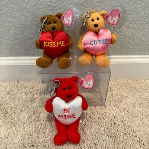 May include: Three plush teddy bear keychains. One brown bear holds a red heart with "KISS ME" in yellow. Another is orange with a pink heart that says "CUTIE". The third is red with a white heart that says "BE MINE".