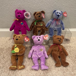 May include: A collection of six plush teddy bears in various colors, including purple, brown, blue, and lavender. Each bear has a red tag with the word "TY" on it. The bears are arranged in a row on a white surface.