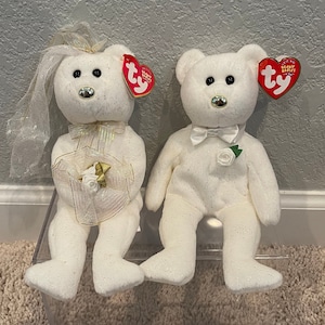 May include: Two white plush teddy bears dressed in wedding attire. The bride bear is wearing a white veil and a gold bow. The groom bear is wearing a white bow tie. Both bears have a red "Ty" tag attached to them.