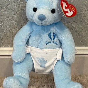 May include: A blue plush teddy bear wearing a white diaper with a blue angel and stars design. The bear has a tag that says "Ty Beanie Babies" and the words "New Baby" are embroidered on the bear's chest.
