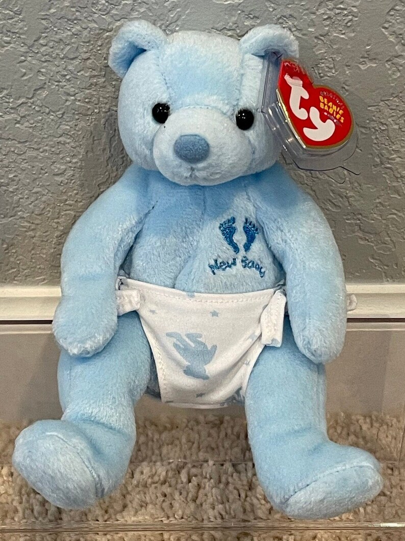 May include: A blue plush teddy bear wearing a white diaper with a blue angel and stars design. The bear has a tag that says "Ty Beanie Babies" and the words "New Baby" are embroidered on the bear's chest.